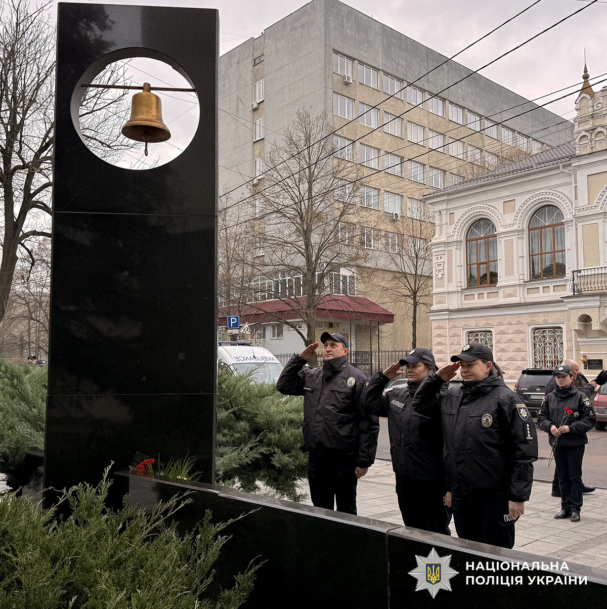 Троє поліцейських у формі стоять біля чорного обеліска з дзвоном та віддають честь. Позаду - міські будівлі. Обеліск прикрашений квітами.