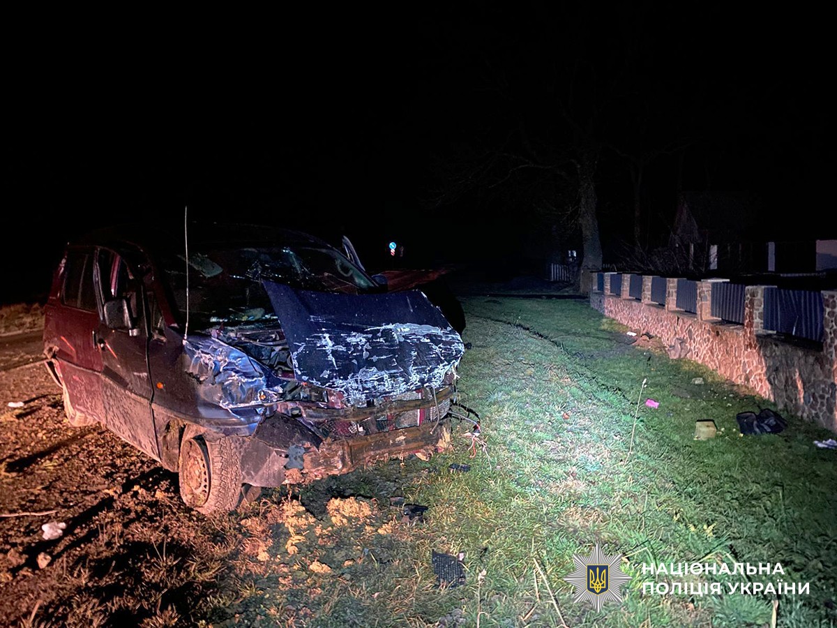 Автомобіль із повністю деформованою передньою частиною на узбіччі біля дороги, навколо сліди удару, уламки та освітлена ділянка місця пригоди.