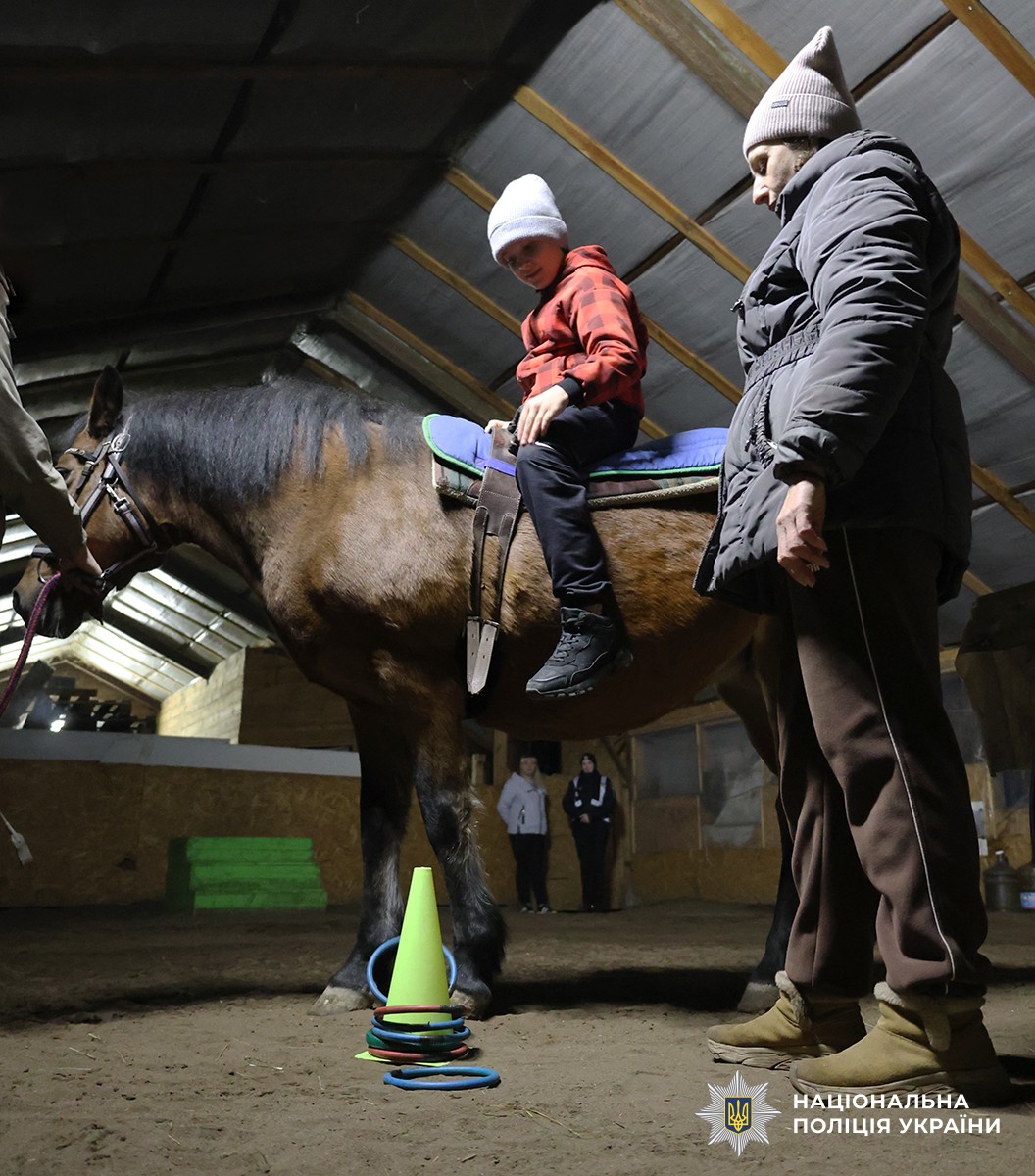 Хлопчик верхи на коні виконує вправу з іграшками. Поруч стоять інструктори. Позаду стоїть поліцейська та мама хлопчика.