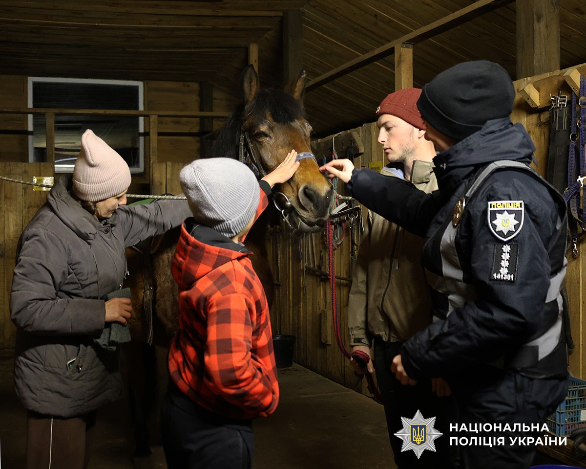 У стайні хлопчик у червоній куртці гладить коня. Поруч із ним стоять поліцейська та інструктори, які спостерігають і допомагають.