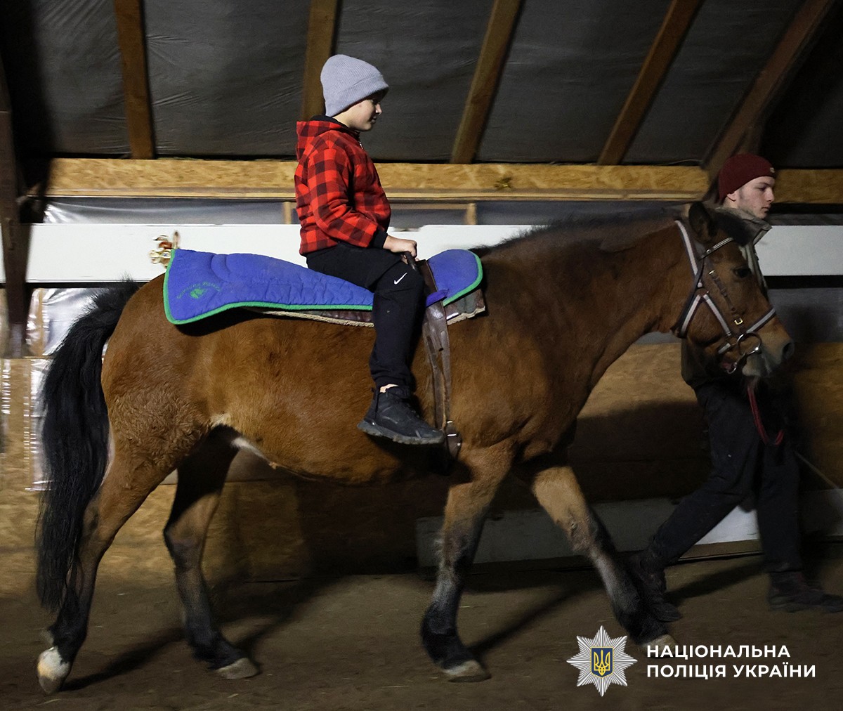 Хлопчик у червоній куртці сидить верхи на коні, поруч іде інструктор.
