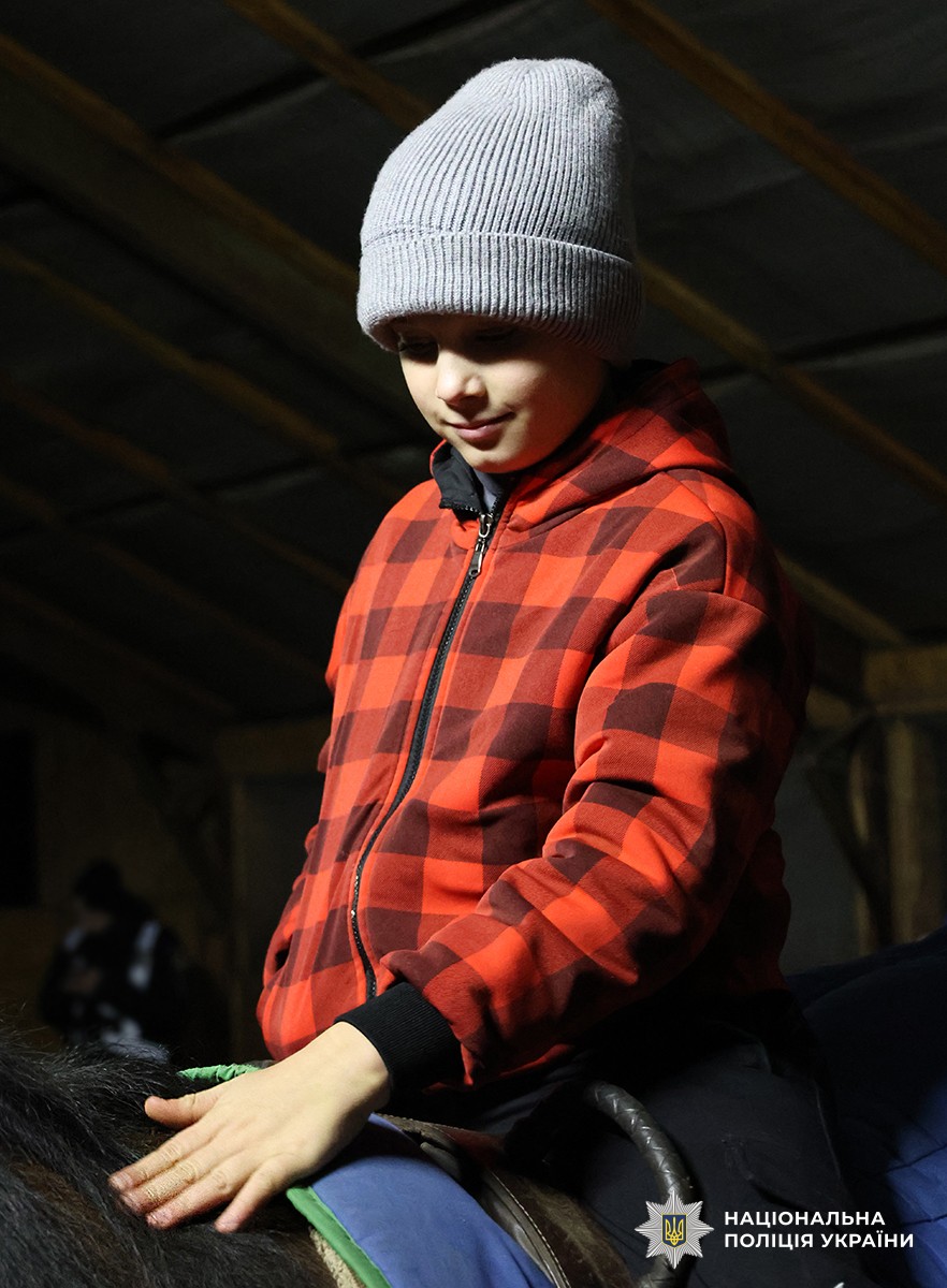 Хлопчик у червоній куртці, сидячи верхи, гладить коня.