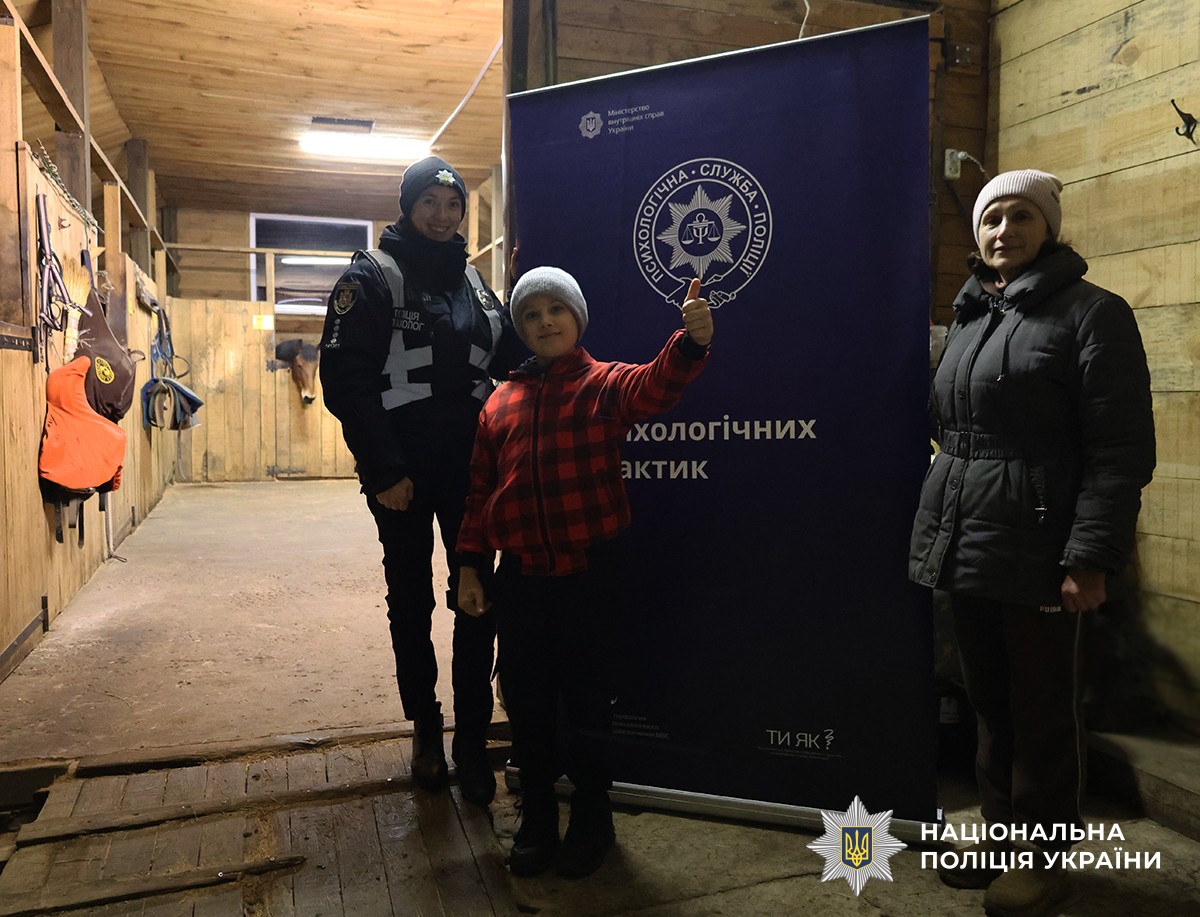 У стайні хлопчик у червоній куртці разом із поліцейською та інструкторкою позують для спільного фото на фоні плаката з емблемою та написом «Пункт психологічних практик».