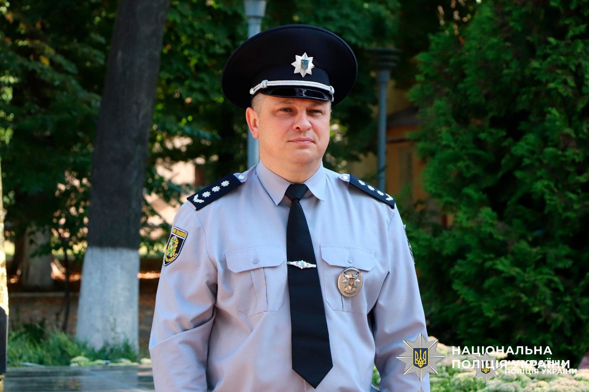 Співробітник Національної поліції України у світло-блакитній сорочці з еполетами та срібним кашкетом стоїть на відкритому повітрі перед зеленим фоном. На формі видно нагрудний знак, що підкреслює офіційний характер зображення.