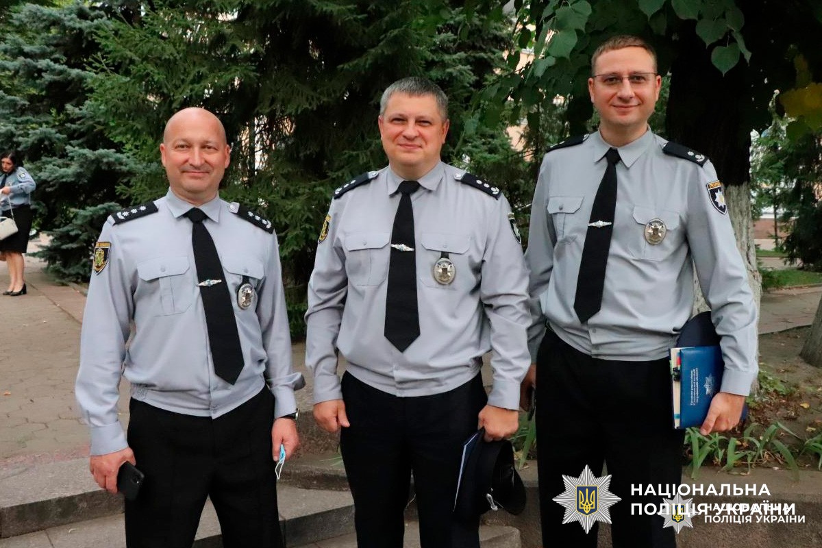 Троє офіцерів Національної поліції України у світло-сірих сорочках стоять на відкритому повітрі.
