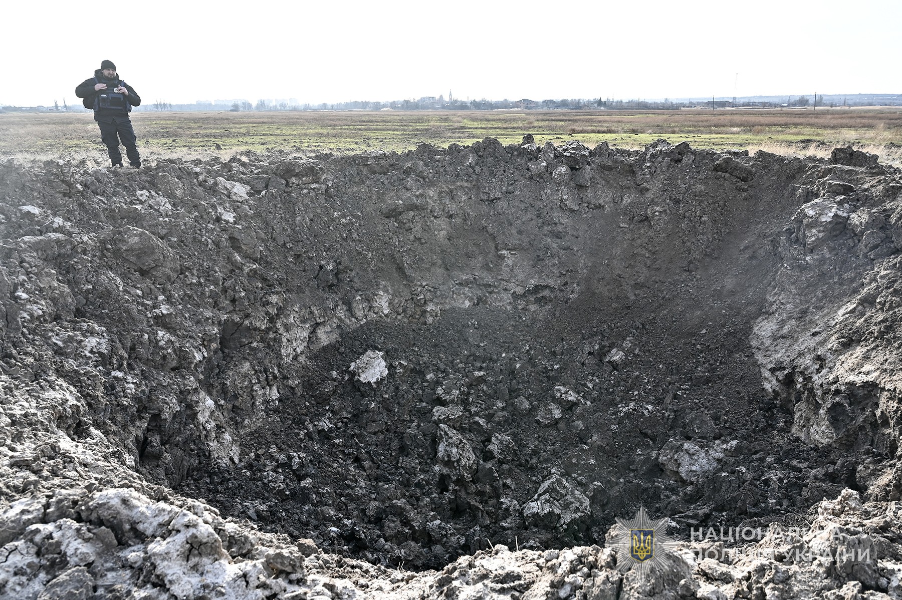 Поліцейський стоїть навпроти вирви від російської авіабомби. Околиці міста Оріхів. Cічень 2025 року.
