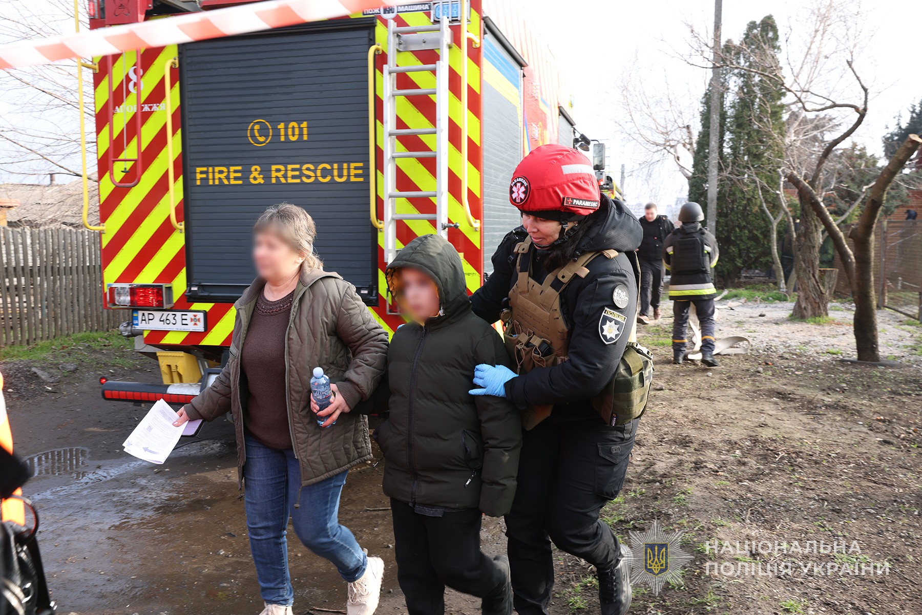 Парамедикиня запорізької поліції виводить матір з дитиною з місця влучання ворожої авіабомби по одній з вулиць обласного центру. Грудень 2025 року.