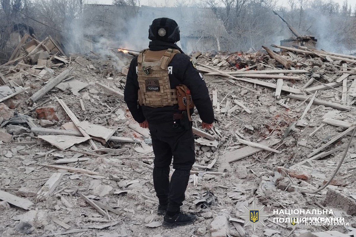 Співробітник Національної поліції України оглядає місце руйнувань після вибуху, серед уламків та диму.