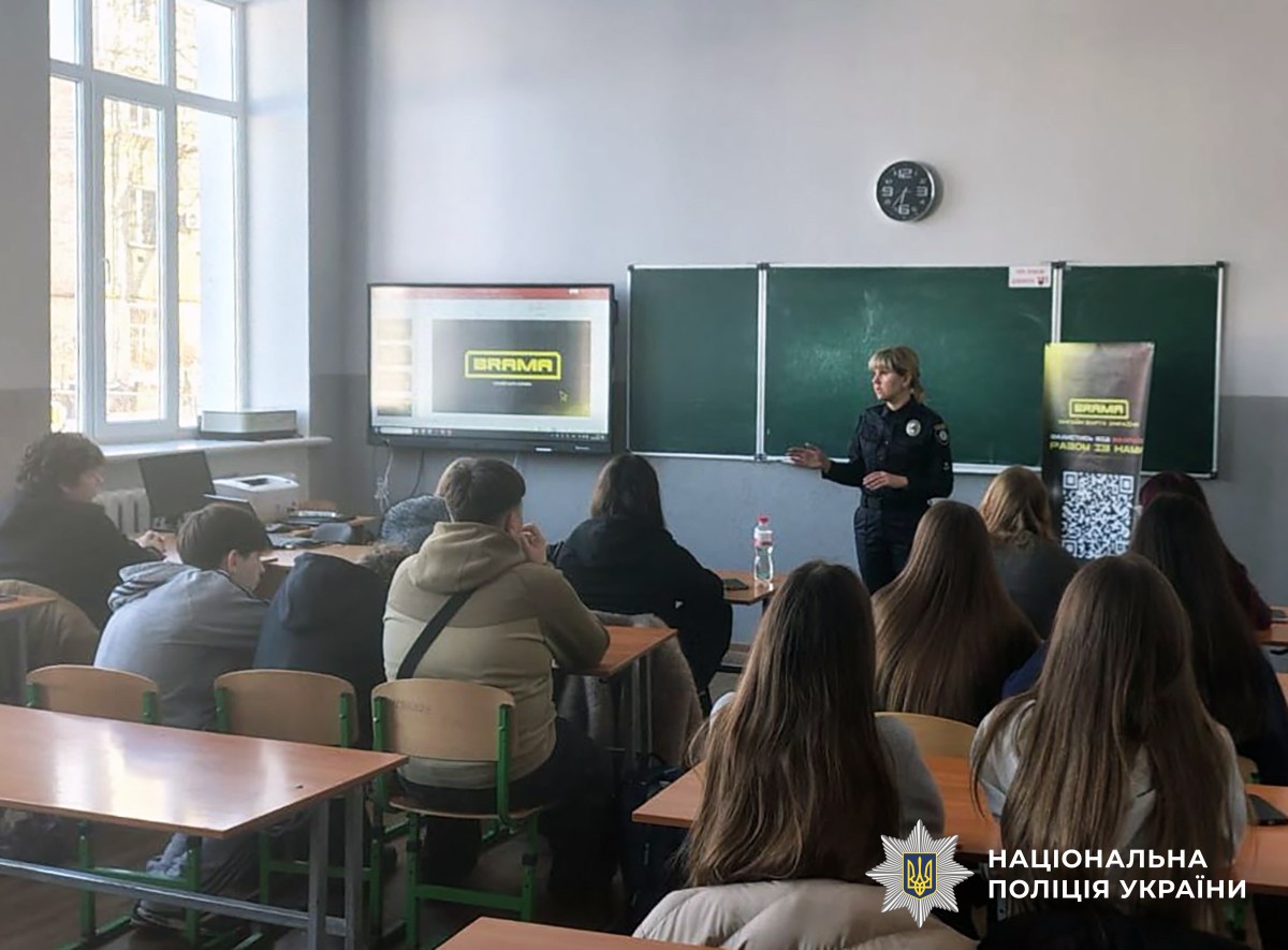 Поліціянтка стоїть біля шкільної дошки перед учнями, які сидять за партами. Поруч розташований екран, на якому написано «BRAMA».