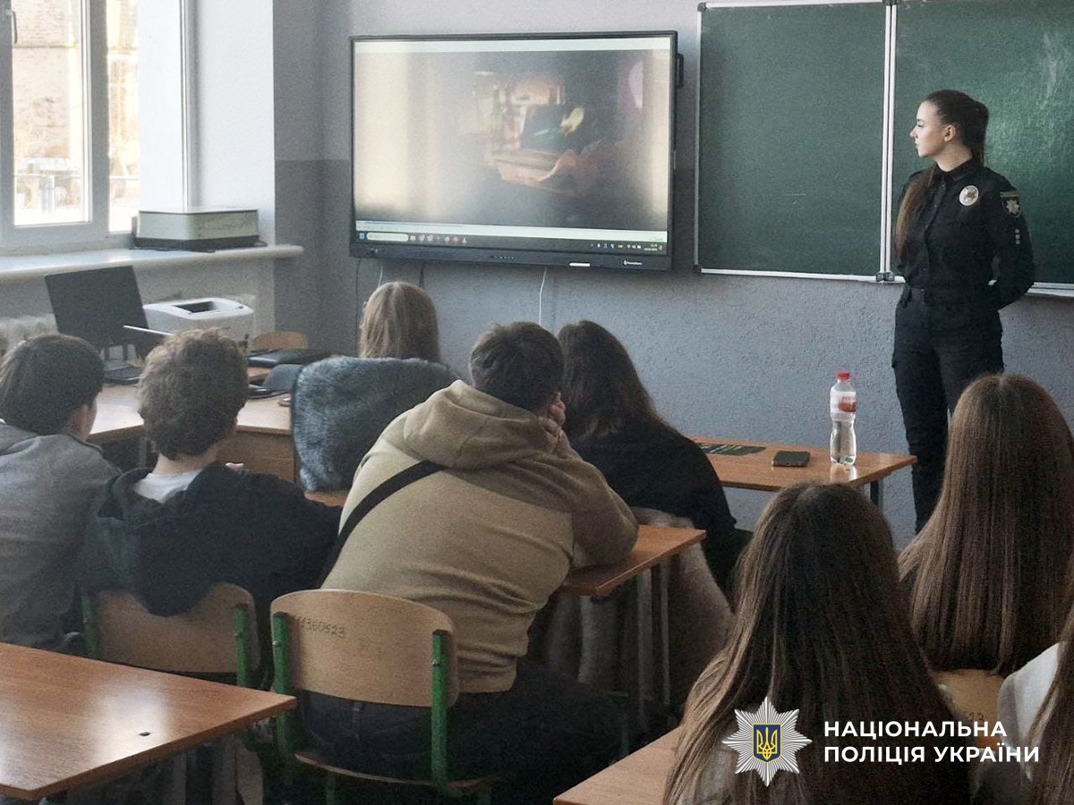 Поліціянтка стоїть біля шкільної дошки перед учнями, які сидять за партами. Учасники переглядають відеоролик на екрані.