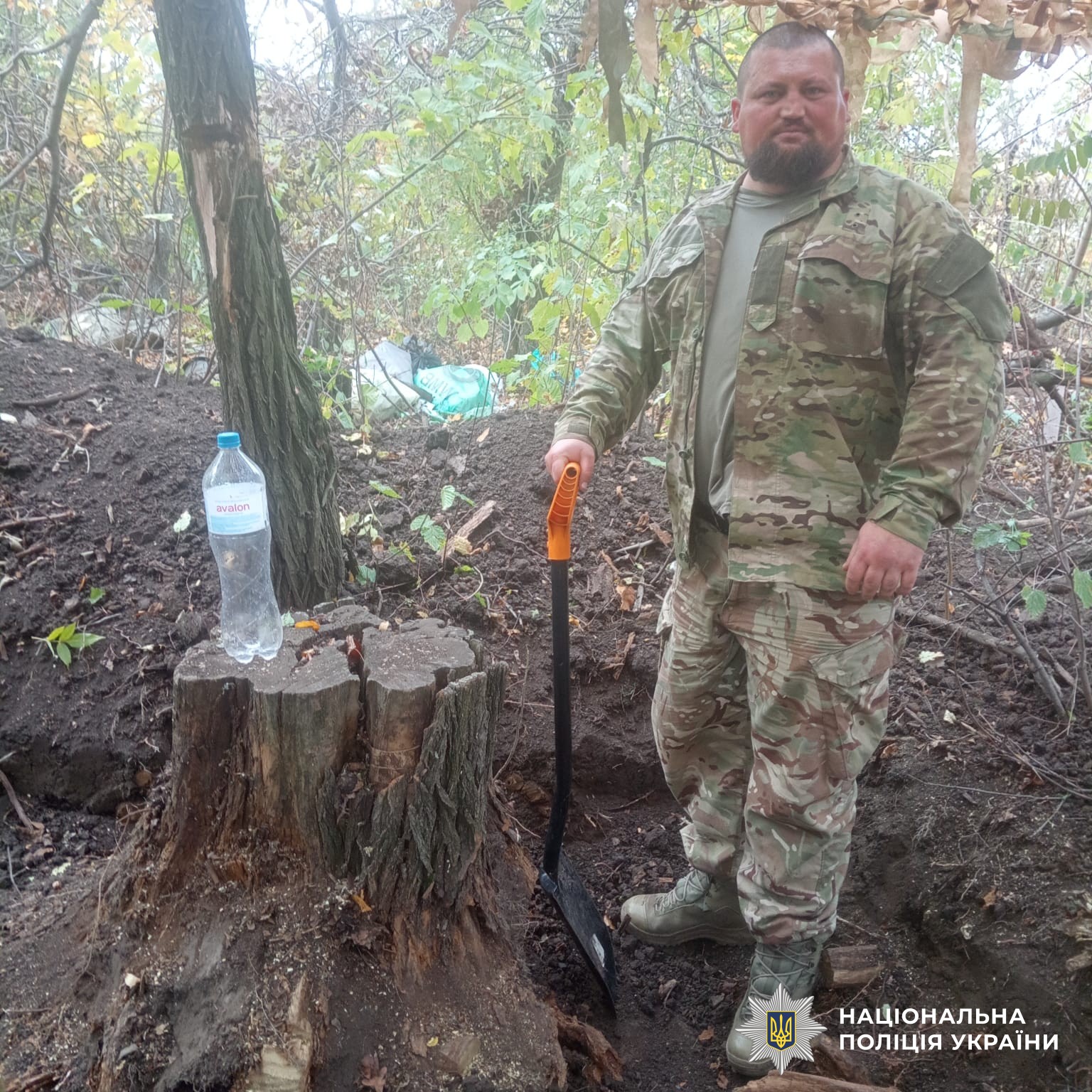Віктор у камуфляжній формі Національної поліції України стоїть у лісовій місцевості біля пенька з пляшкою води та тримає невелику лопату