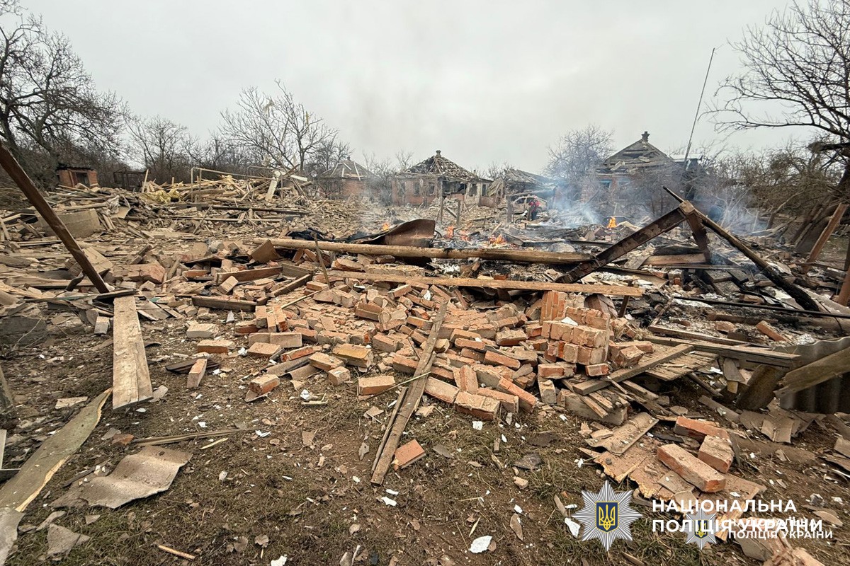 На передньому плані уламки цегли та деревини, у глибині пошкоджені будівлі з осередками пожежі.