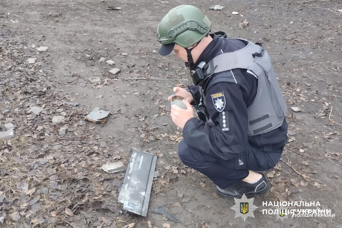 Поліцейський у захисному спорядженні оглядає металевий предмет серед уламків після вибуху.