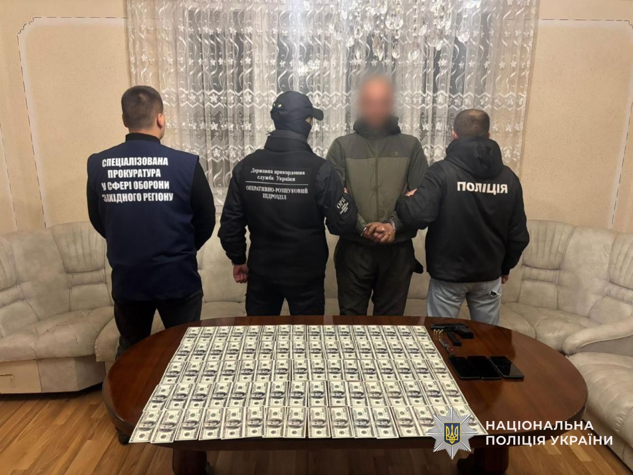 Правоохоронці тримають підозрюваного у переправленні через кордон. Попереду них — стіл, на якому розкладена готівка, вилучена у фігуранта