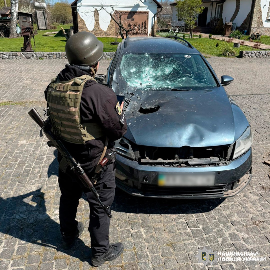 Cпівробітник Національної поліції України у бронежилеті та касці, який оглядає пошкоджений автомобіль. Машина має значні сліди ураження: розбите лобове скло з численними отворами, пробиту капотну частину та пошкоджену решітку радіатора.